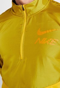 Chaqueta amarilla con media cremallera y un patrón texturizado, con el logo de Nike en naranja. Presenta un cuello alto y un frontal con cremallera. Tejido ligero.