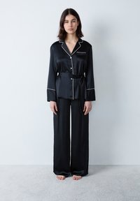 Zwarte satijnen pyjama set met een lange mouwen top met witte bies, knopen aan de voorkant en bijpassende wijde broek. Geen patronen of accenten.