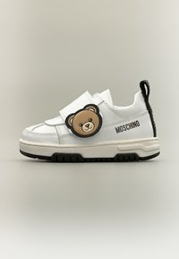 Λευκό αθλητικό παπούτσι Moschino με μαύρη σόλα, ιμάντα βέλκρο και καφέ διακοσμητικό πρόσωπο αρκουδάκι στο πλάι.