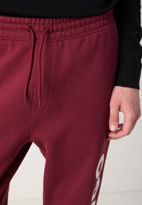 Burgunderfarbene Jogginghose mit einem elastischen Bund und Kordelzug. Sie verfügt über eine kleine seitliche Tasche und ein weißes Logodetail am Bein.