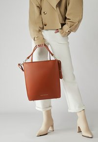 Hnědá kožená taška přes rameno se čtvercovým tvarem, dvojitými úchyty a odnímatelným ramenním popruhem. Má hladkou texturu a detaily s logem.