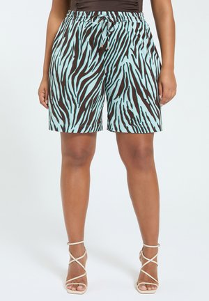 Frau trägt midilange Shorts mit Zebramuster in Türkis und Schwarz mit Kordelzugbund und beigen Riemchen-Sandalen mit hohen Absätzen.