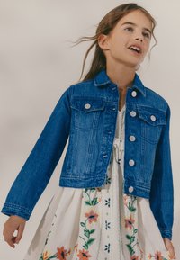 Fiatal lány kék farmerdzsekit visel fehér ruha felett, amely színes virágos hímzéssel díszített, áll és oldalra néz.