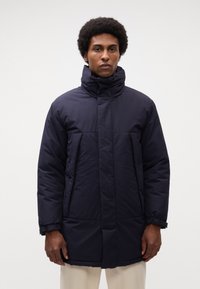 Cappotto imbottito blu navy con colletto alto, maniche lunghe e tasche frontali. Texture liscia con una finitura leggermente lucida.