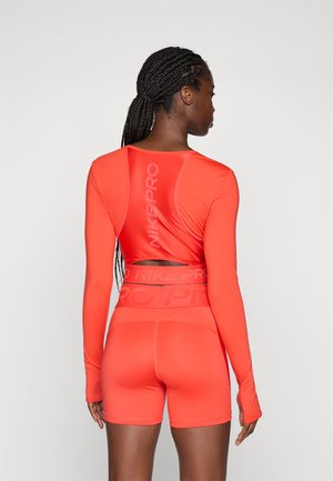 Woman with braided hair wearing a bright orange long-sleeve crop top and matching high-waisted shorts with "Nike Pro" branding.