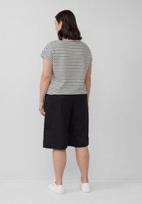 Vrouw met schouderlang donker haar, gekleed in een wit-zwarte gestreepte top, zwarte knielange shorts en witte sneakers, staande met het gezicht naar een muur.