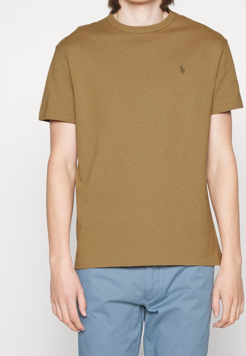 Hnědé bavlněné tričko s kulatým výstřihem a krátkými rukávy, na hrudi s malým logem. Nosené s modrými kalhotami.