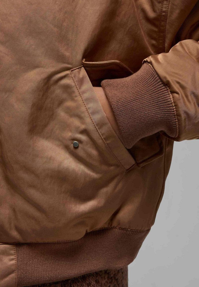 Veste bombardier marron avec une texture lisse et brillante, des poignets côtelés et une poche latérale dotée d'une fermeture à pression métallique.