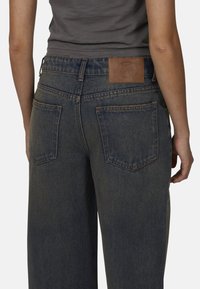 Jeans en denim bleu foncé avec une taille haute, comportant deux poches arrière et une étiquette de marque en cuir marron cousue sur la ceinture.