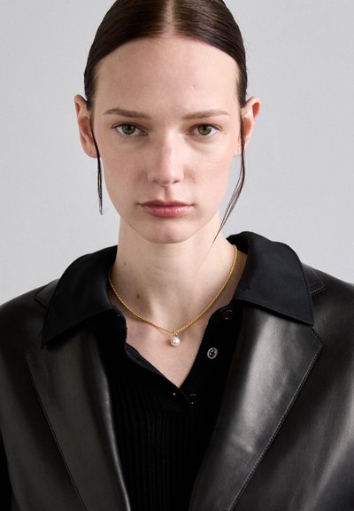 Collarín de oro trenzado con una sola perla blanca, llevado sobre una camisa negra con cuello y una chaqueta de cuero. Texturas suaves y colores contrastantes.