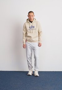 Lys beige hettegenser med logo, grå joggebukser og hvite sneakers. Hettegenseren har en frontlomme og snøring. Enkel, uformell design.