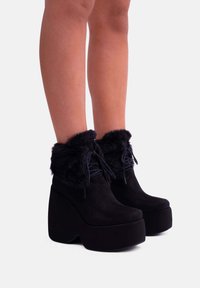 Stivali platform in suede nera con risvolto in finta pelliccia, punta rotonda e dettagli con lacci. La suola è spessa e curva per aggiungere altezza.