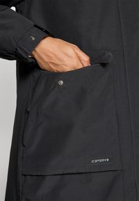 Hand gedeeltelijk in de grote voorzak van een zwarte Icepeak jas met drukknoopsluiting en een ritsdetail.