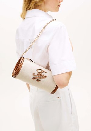 Femme portant une tenue blanche porte un sac à main cylindrique beige avec un bord marron et une bandoulière chaîne dorée sur l'épaule.