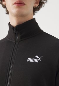 Melnā virsjakas ar rāvējslēdzēju, izgatavota no mīksta auduma, ar augstu apkakli un baltu izšuvumu Puma logo uz kreisā krūšu sāna.