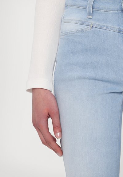 Lys blå denimjeans med en glatt tekstur, høy midje-design og subtil søm. En hvit langermet topp med ribbestrikket detalj delvis synlig.