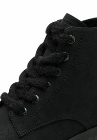 Bottine noire en cuir avec de gros lacets noirs duveteux passés dans des œillets argentés et une couture détaillée sur le côté.