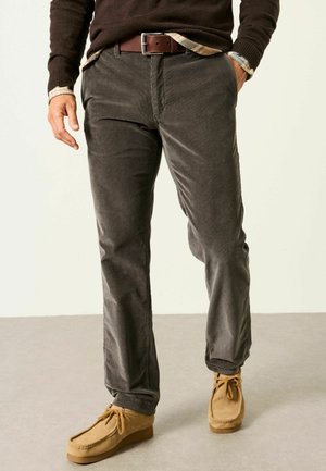 Mann trägt dunkelbraune Cordhose, braune Wildleder-Schnürschuhe, einen braunen Pullover und ein kariertes Hemd mit den Händen in den Taschen.