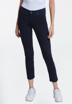 Mujer vestida con jeans ajustados cortos de color azul oscuro, zapatillas blancas y una blusa sin mangas de color azul claro, de pie contra un fondo blanco.