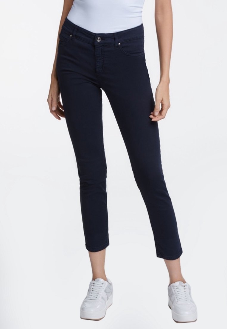 Žena v tmavě modrých skinny cropped džínách, bílých teniskách a svetru bez rukávů světle modré barvy, stojící na bílém pozadí.