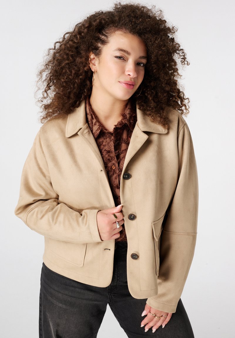 Jonge vrouw met krullend haar, gekleed in een beige jasje met knopen, een bruine blouse met structuur, zwarte broek en gouden oorringen, tegen een witte achtergrond.