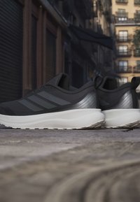 Pair of black and gray athletic shoes with white soles placed on a cobblestone street with urban buildings in the background.