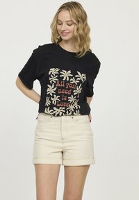 T-shirt en coton noir à manches courtes avec un imprimé floral blanc et un texte rouge indiquant "Tout ce dont vous avez besoin, c'est d'amour", associé à un short beige.