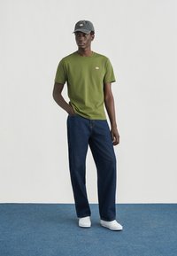 Πράσινο βαμβακερό μπλουζάκι με μικρό λογότυπο στο στήθος, σε συνδυασμό με σκούρο μπλε τζιν παντελόνι και λευκά αθλητικά παπούτσια, φορεμένο με γκρι καπέλο.