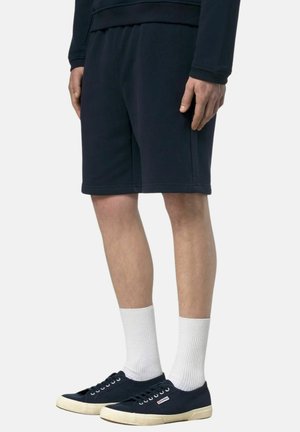 Uomo che indossa pantaloncini neri, calzini bianchi alti e scarpe da ginnastica nere basse, in piedi davanti a uno sfondo uniforme.