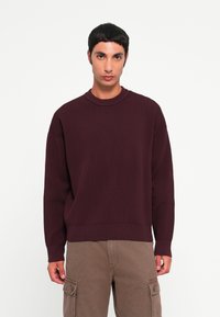 Pull en maille côtelée bordeaux à col rond et épaules tombantes, associé à un pantalon cargo marron avec poches latérales.