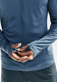 Granatowa sportowa kurtka z długim rękawem, pełnym zamkiem, otworami na kciuki i gładką fakturą, zestawiona z luźnymi czarnymi szortami.