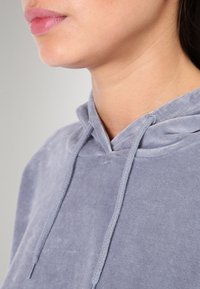 Šedá sametová mikina s kapucí a stahovací šňůrkou. Vyznačuje se měkkou, hladkou texturou a uvolněným střihem. Viditelné švy na kapuci a oblasti krku.