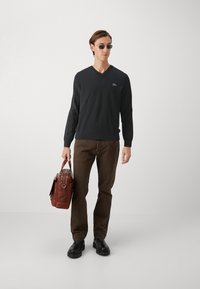 Pulover negru cu decolteu în V, pantaloni maro, cizme negre, ținând o geantă din piele maro. Accesoriile includ ochelari de soare închiși la culoare. Design simplu și funcțional.