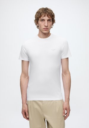 Junger Mann mit hellbraunen lockigen Haaren, der ein schlichtes weißes kurzärmeliges T-Shirt und beige Hosen trägt, vor einem weißen Hintergrund steht.