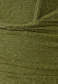 Olivově zelená texturovaná látka s vlnitým vzorem. Materiál vypadá lehký a síťový, s nabranými detaily a hladkým zipovým uzávěrem.