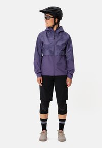 Lila wasserdichte Jacke mit hohem Kragen und Frontreißverschluss, kombiniert mit schwarzen Shorts und schützenden Knieschonern. Graue Schuhe runden das Outfit ab.