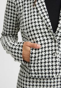 Bleđi sivi sako s uzorkom jagnjeće glave (houndstooth) u crnoj i bijeloj boji, s jednim gumbom, reverima s urezom i džepom s lijeve strane.