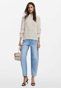 Maglione a maglia color crema con macchie colorate, vestibilità rilassata, polsini a coste, abbinato a jeans dritti blu chiaro e tacchi neri. Porta una borsa beige.