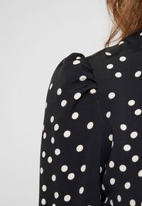 Svart blus med vita prickar, med puffad axeldetalj och en mjuk textur. Närbild på ärmdesign och mönster.