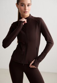 Mujer que lleva una chaqueta ajustada marrón oscuro con cremallera y leggings a juego, posando con una mano cerca del cuello y la otra en la cadera.