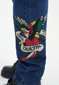 Jeans de mezclilla azules con un diseño colorido de cristales de imitación que presenta un corazón, un águila y una cinta con las palabras "Verdadera Muerte" en blanco y negro.