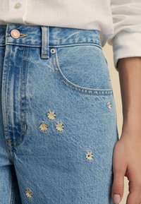 Vaqueros de denim de color azul claro con bordados florales de pétalos blancos y centros amarillos, detalles desgastados y un diseño clásico de cinco bolsillos.