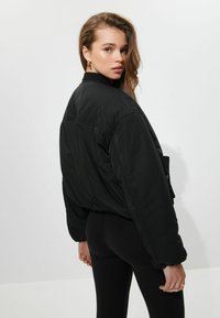 Jeune femme avec des cheveux lâches portant un bomber noir surdimensionné et des leggings noirs, se retournant vers l'arrière devant un fond uni.