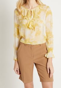 Ljusgul blus med volang i halsen och långa ärmar, ihop med bruna skräddarsydda shorts. Blusen har ett akvarellmönster.