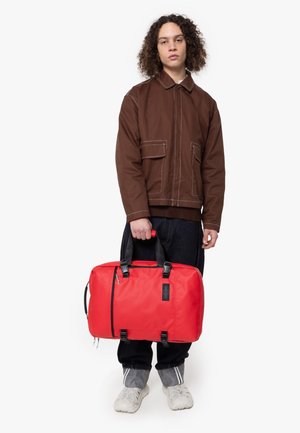 Sac de sport rouge avec des accents noirs, de forme rectangulaire, doté de poignées supérieures et de doubles fermetures éclair. Le modèle porte une veste marron et un jean foncé.