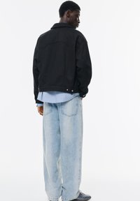 Jachetă din denim neagră cu o croială lejeră, având guler, închidere cu nasturi și detalii pe spate, stratificată peste o cămașă dungată de culoare albastră deschisă și blugi deschiși.