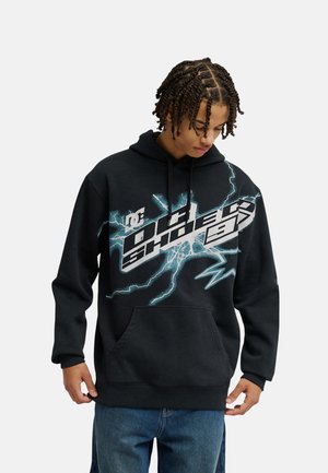 Junge Person mit geflochtenen Haaren blickt nach unten, trägt einen schwarzen DC Shoes Hoodie mit auffälligem Blitzmotiv und blaue Jeans vor einfarbigem Hintergrund.