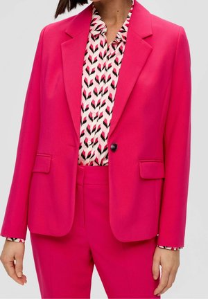 Kvinde iført en knaldrød dragt med en enkeltskaldet blazer og mønstret bluse i hvide, sorte og pink nuancer.