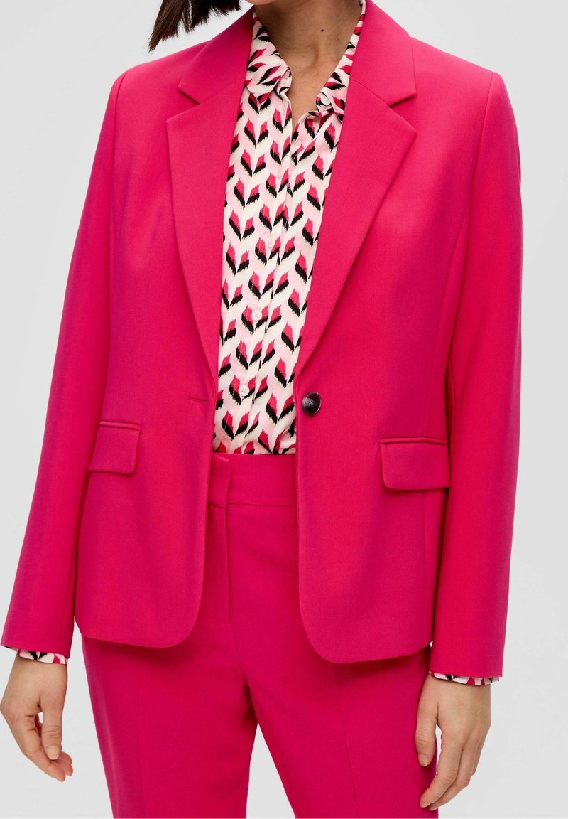 Femme portant un costume rose vif avec un blazer à un bouton et un chemisier à motifs blancs, noirs et roses.