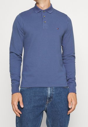 Mand iført en blå poloshirt med lange ærmer og knapper samt blue jeans, stående mod en ensartet hvid baggrund.